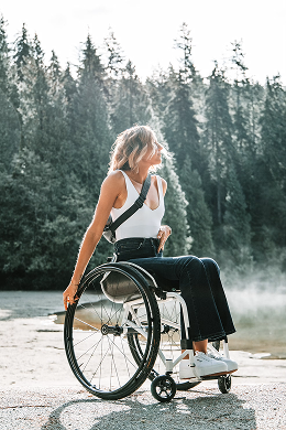Elyse smiling in her wheelchair by a lake, wearing a white tank top and dark jeans