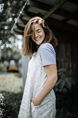 Della, an occupational therapist, smiling outdoors in a white printed pinafore over a blue t-shirt
